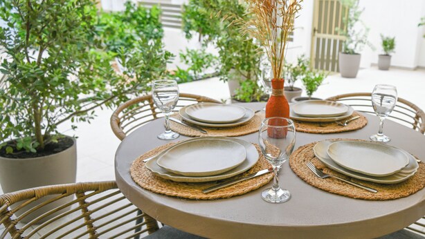 A round table set for four with plates, cutlery, and glasses, surrounded by wicker chairs and potted plants. - Vivis Hub (Kefalonia)