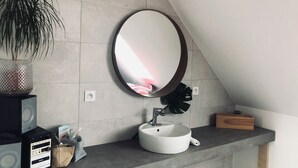 Un baño con un lavamanos blanco, un espejo redondo, una planta en maceta y un altavoz.