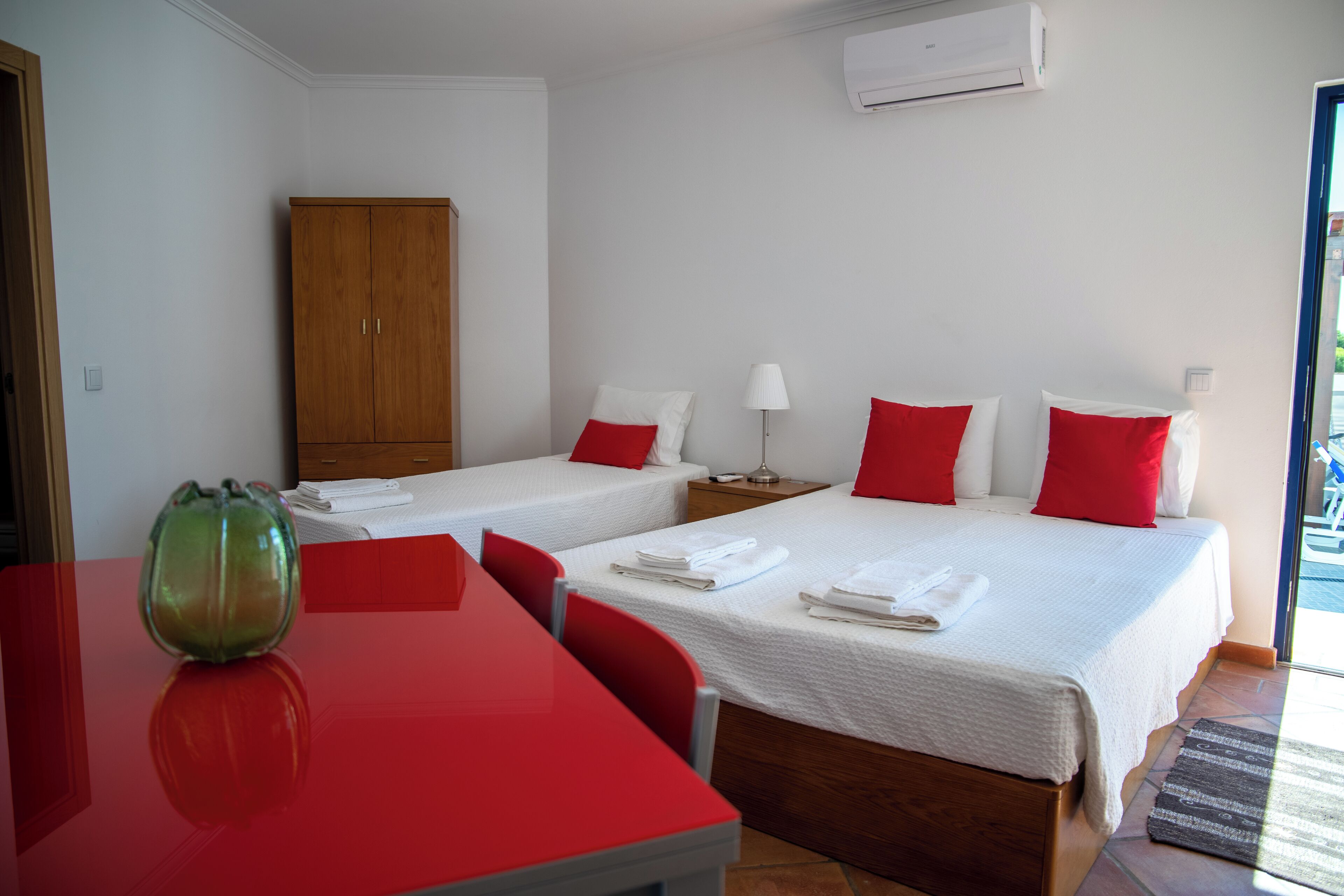 A hotel room with two beds, a wooden wardrobe, a red table, and a glass vase.