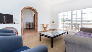 A living room with a TV, seating, a glass coffee table, a brick archway, and a balcony with outdoor furniture. - Monte da Eirinha (Mertola)