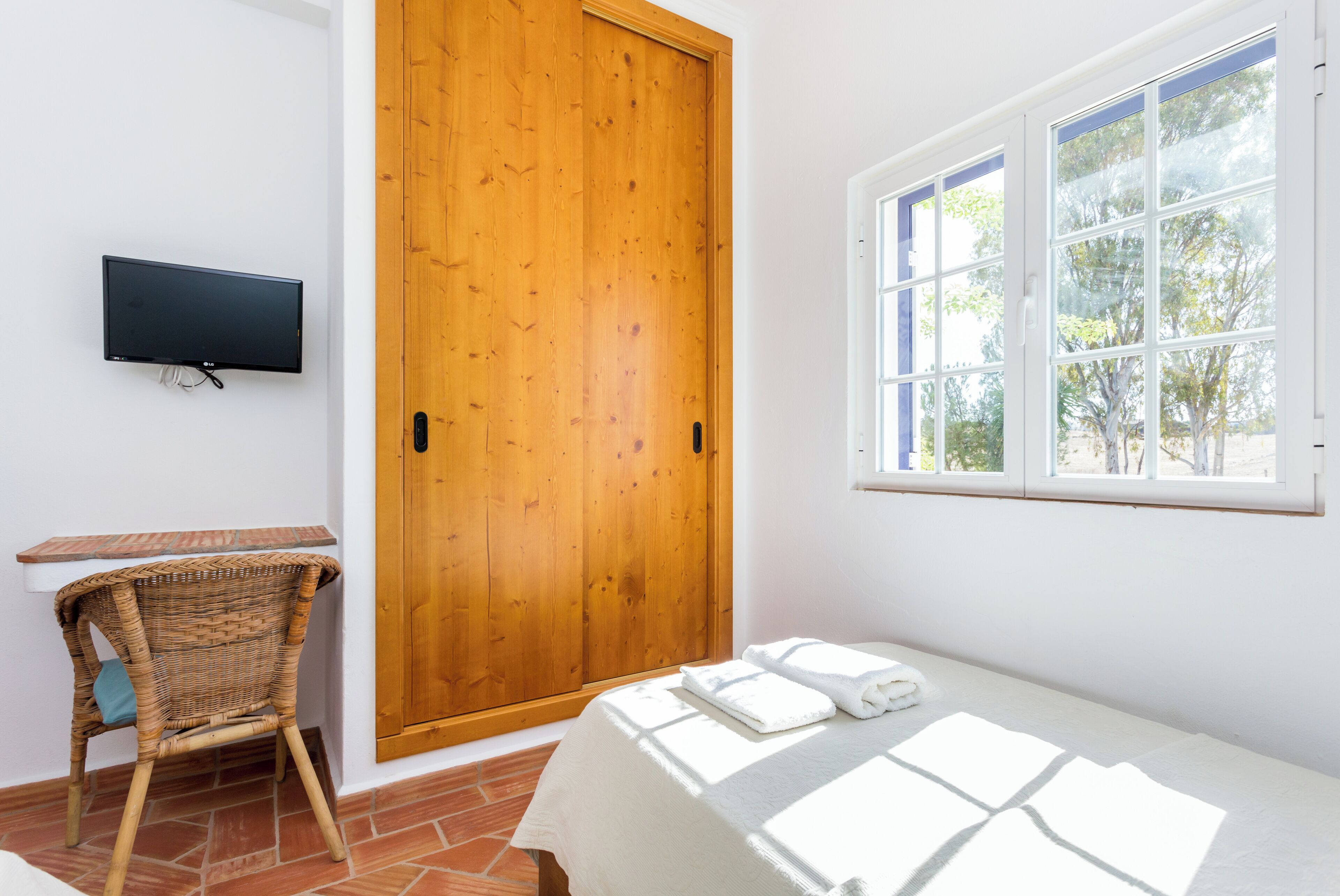 Une chambre avec un lit, un téléviseur fixé au mur, une porte en bois et une fenêtre donnant sur des arbres.