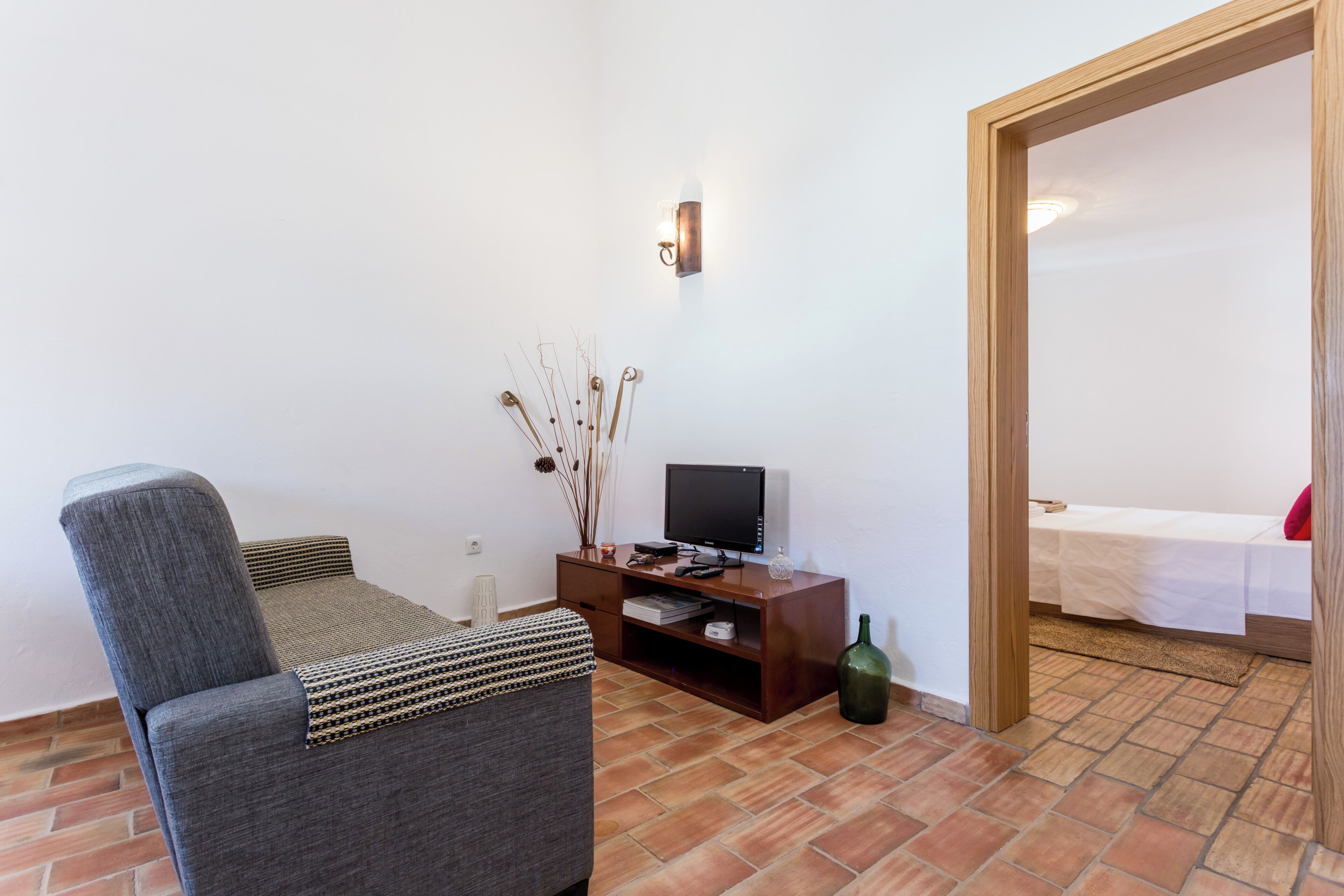 A small, furnished room with a grey sofa, a wooden TV stand, a TV, and a bed with a white bedspread and red pillow.