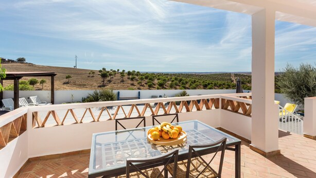 A terrace with a glass table and chairs, a bowl of fruit, and a view of a landscape with trees and a distant hill. - Monte da Eirinha (Mertola)