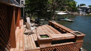 Una terraza de madera con mobiliario exterior, un pequeño bote amarrado y vistas al agua y árboles.