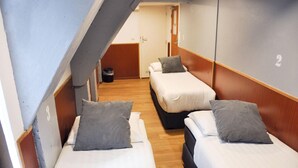 Un pasillo estrecho con tres camas alineadas contra la pared, cada una con almohada y colchón blanco. Se ve una puerta al final del pasillo.