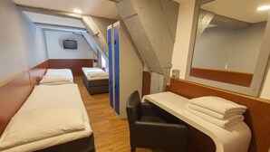 Habitación compacta con dos camas individuales, un escritorio pequeño con una silla y un televisor en la pared.