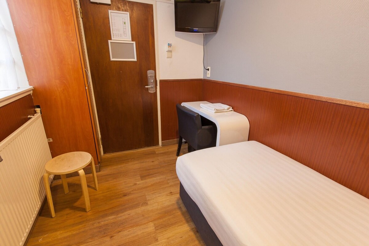 A small, neatly arranged room with a single bed, a chair, a small table, and a television mounted on the wall.