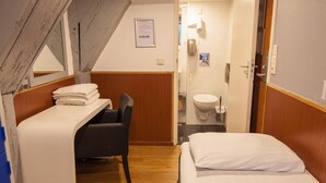 Habitación de hotel compacta con literas, un escritorio con silla, baño pequeño con inodoro y lavamanos, y techo de madera.