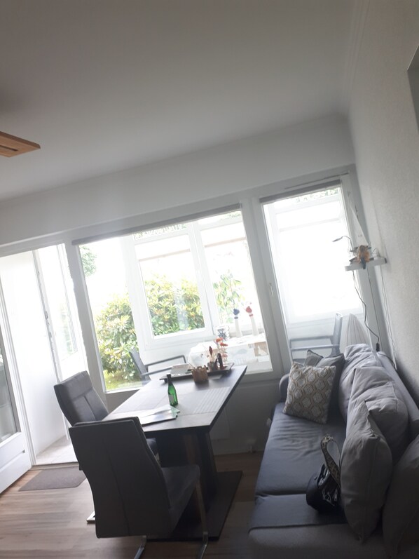 A modern living room with a sofa, a dining table, and a chair. There are large windows providing natural light and a view of greenery outside. - Fördetraum,1. Reihe, 20 m zum Strand, Meerblick, Frisch Renoviert, Loggia, EG (Laboe)