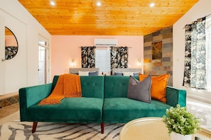 A green sofa with a throw blanket, a small table with a plant, and a wall-mounted air conditioner. - Vegas 4BR: Poker • Wet Bar • Parking (Las Vegas)