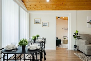 A dining area with a wooden ceiling, white walls, and a view into another room with a sofa and a plant.