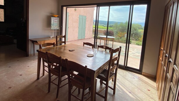 Un espace repas avec une grande table et des chaises, un lustre, une vue à travers une porte vitrée et un tableau accroché au mur.