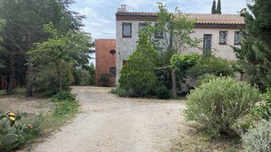 Un chemin de gravier mène à un bâtiment doté d’un toit en tuiles et de murs en pierre, qui est entouré de divers arbres et arbustes.