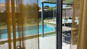 A view through a window of a backyard with a swimming pool, outdoor furniture, and a covered patio area. - Villa Mapokia 2 Climatisée Avec Piscine Chauffée Privative Proche Uzès (st marcel de careiret)