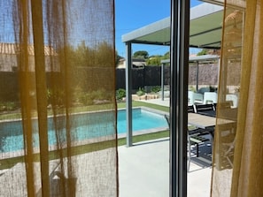 A view through a window of a backyard with a swimming pool, outdoor furniture, and a covered patio area. - Villa Mapokia 2 Climatisée Avec Piscine Chauffée Privative Proche Uzès (st marcel de careiret)
