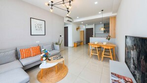 A modern living room with a grey sofa, orange patterned pillows, a coffee table, and a dining area with a wooden table and chairs. - The Tresor Apartment Quan 4 (Ho Chi Minh City)