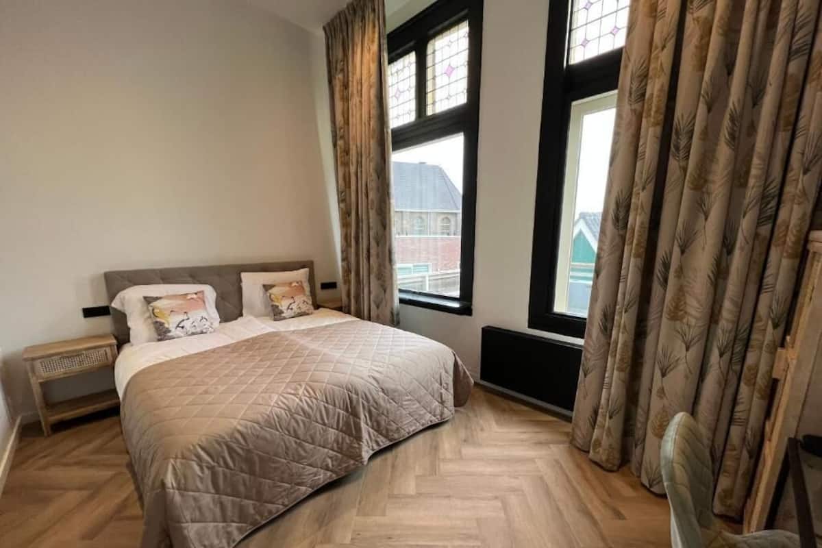 Une chambre à coucher avec un grand lit, une table de chevet, une chaise et une fenêtre avec des rideaux.
