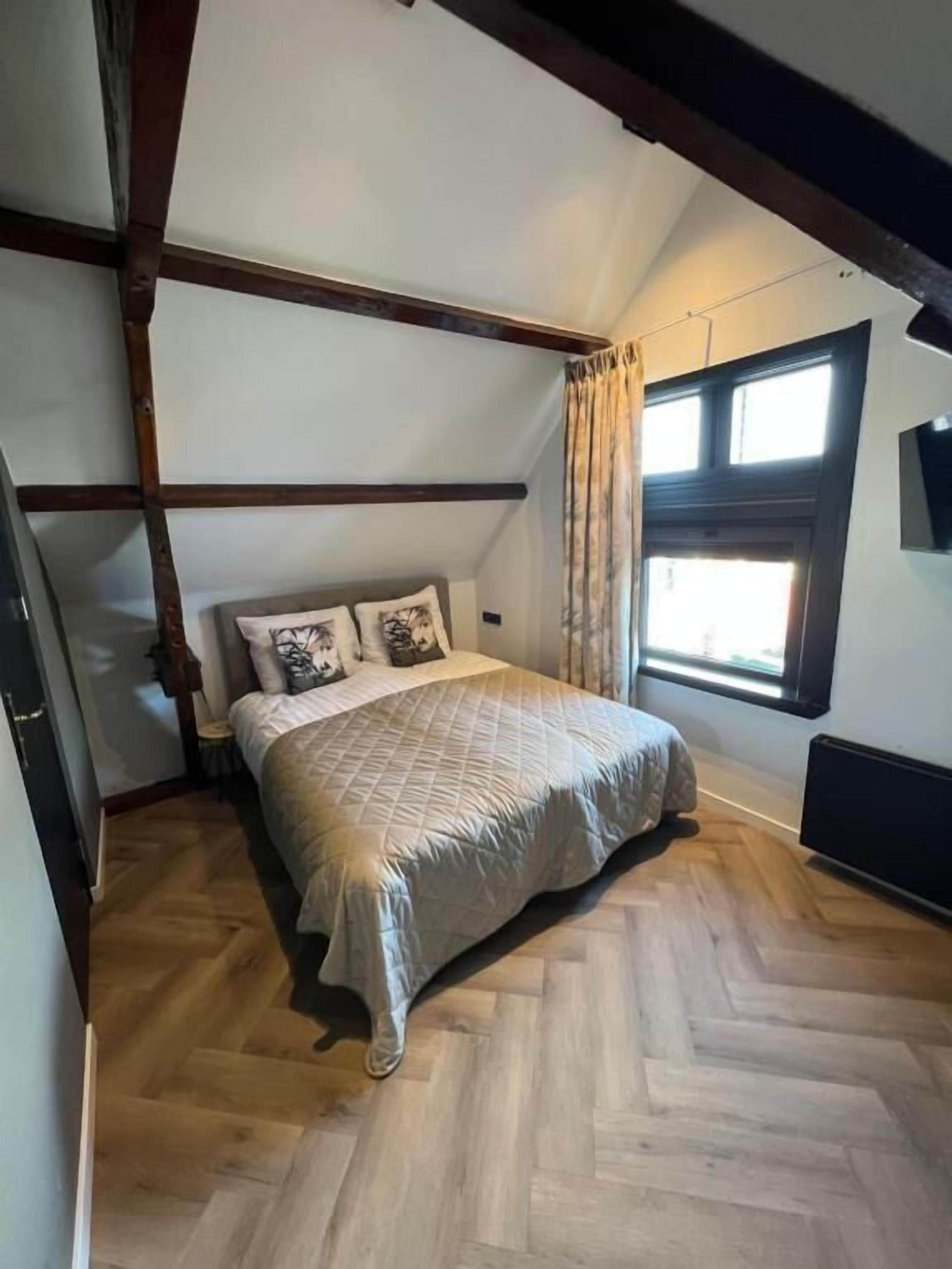 A bedroom with a wooden floor, a bed with a quilted cover, a window with curtains, and exposed wooden beams.