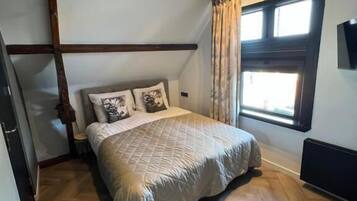A bedroom with a wooden floor, a bed with a quilted cover, a window with curtains, and exposed wooden beams.