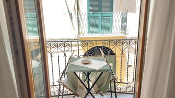 Ένα μπαλκόνι με ένα τραπέζι και καρέκλες, ένα μπολ πάνω στο τραπέζι και θέα σε ένα κτίριο με πράσινες παραθυρόφυλλες.