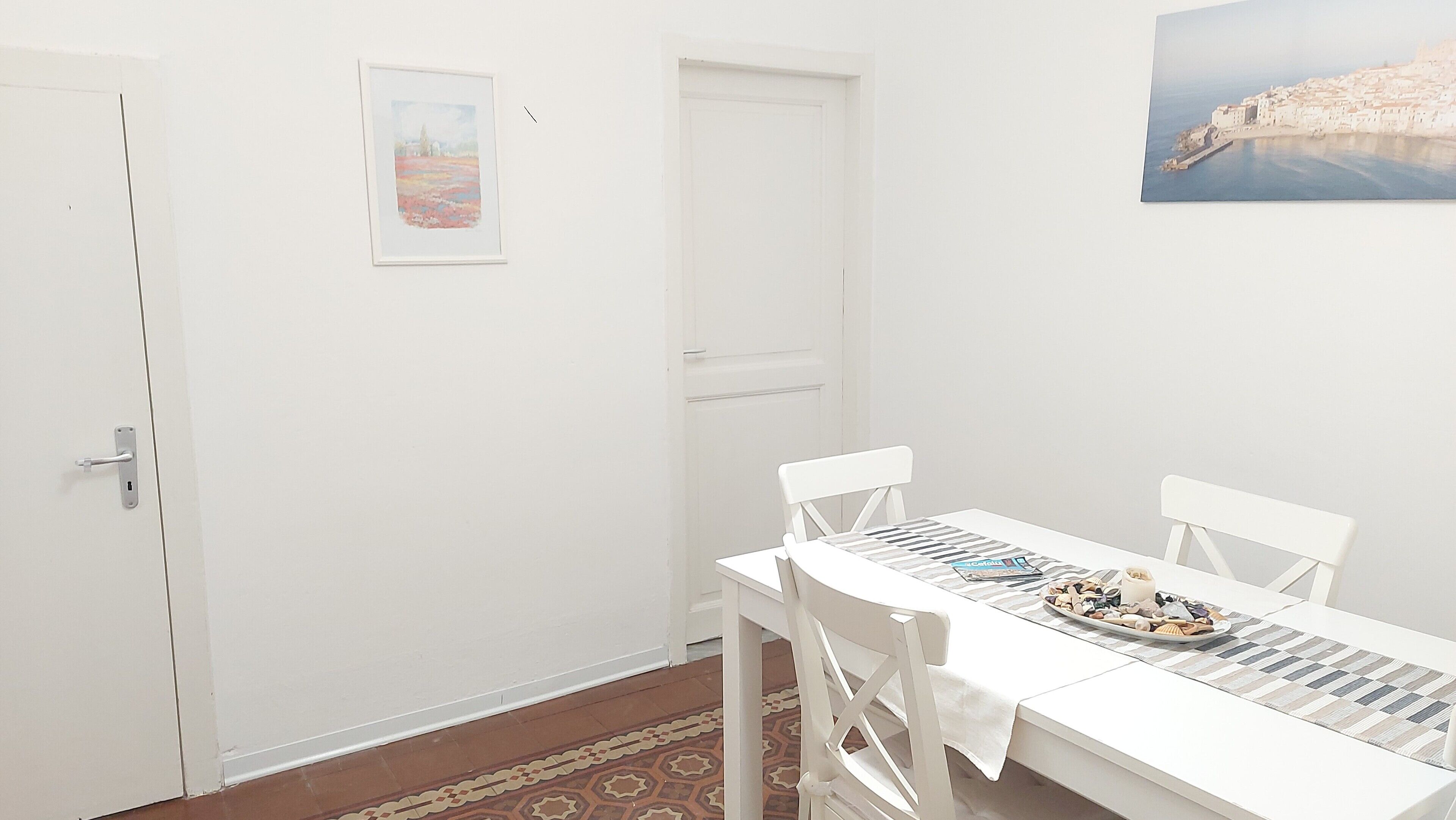 Une pièce blanche avec une table à manger dressée pour deux personnes, une porte et un tableau encadré accroché au mur.