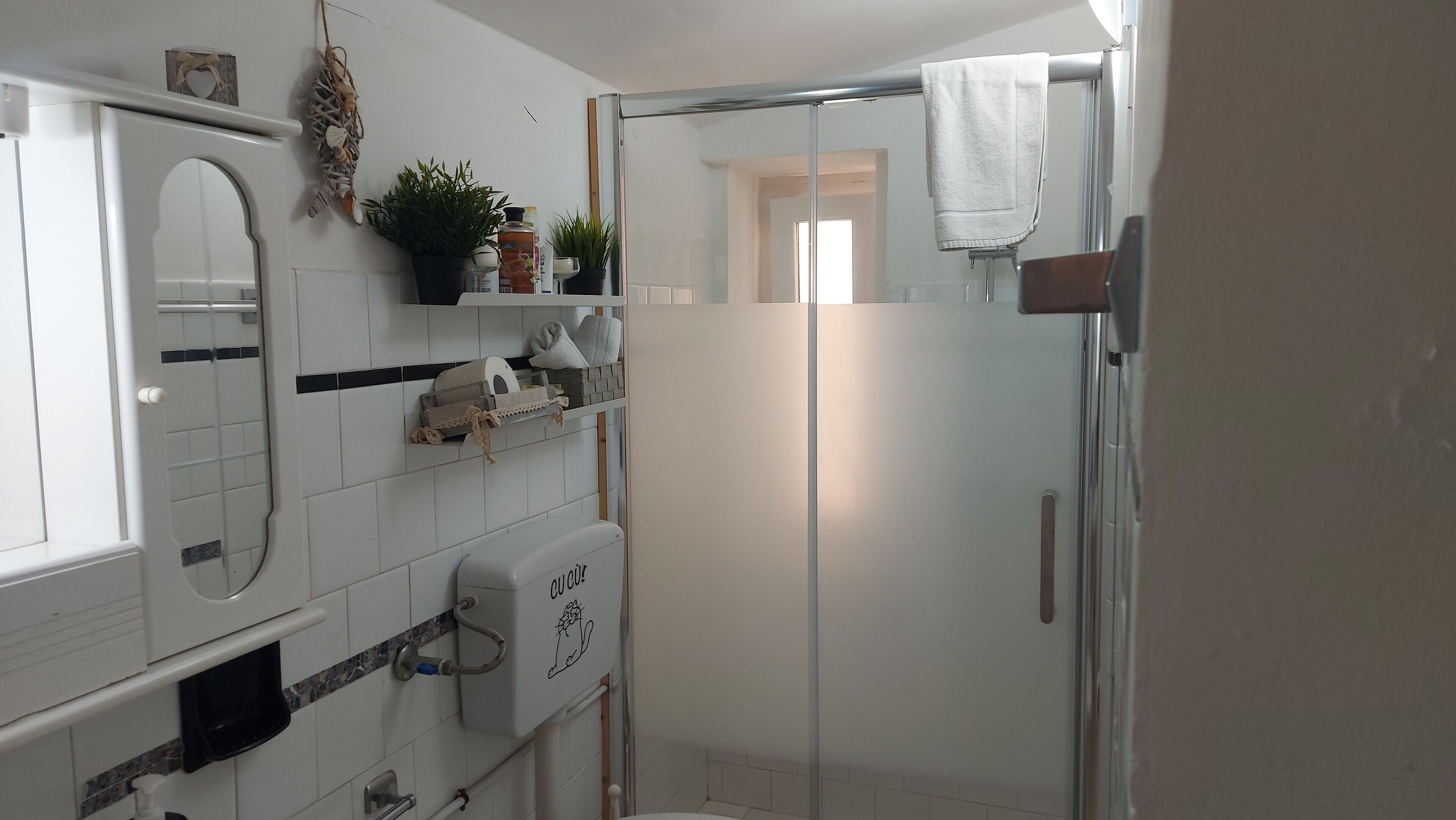 A bathroom with white tiles, a glass shower enclosure, a towel hanging on the shower door, and a mirror reflecting the room.