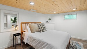 Una camera da letto con soffitto in legno, un letto con biancheria bianca, un tavolino con una lampada e una finestra.