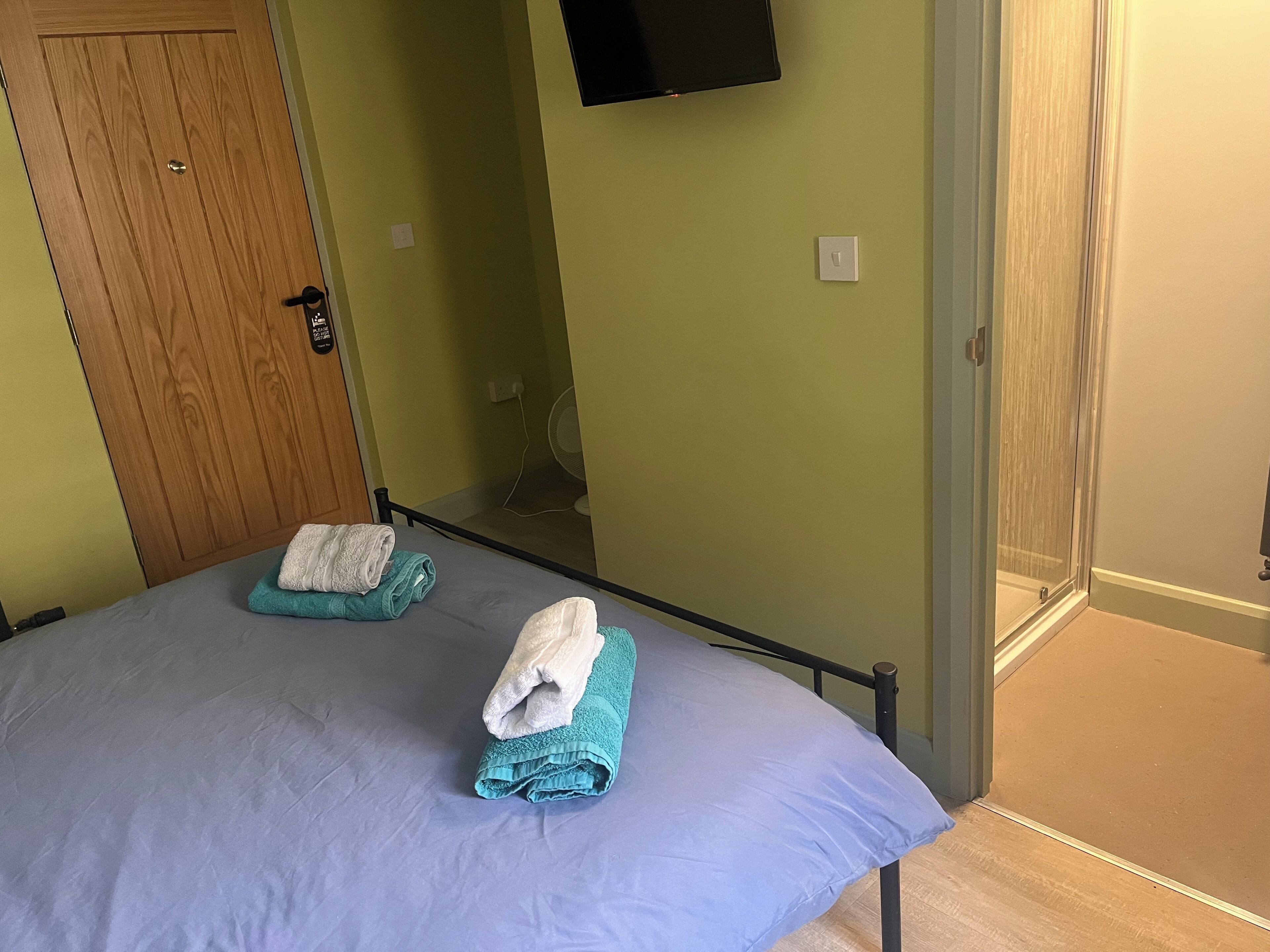 A room with two beds, a wooden door, a TV mounted on the wall, and a small fan.