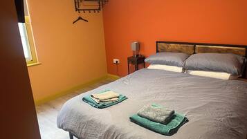A bedroom with a bed, two pillows, a nightstand with a lamp, and a wall-mounted shelf with a hanger.
