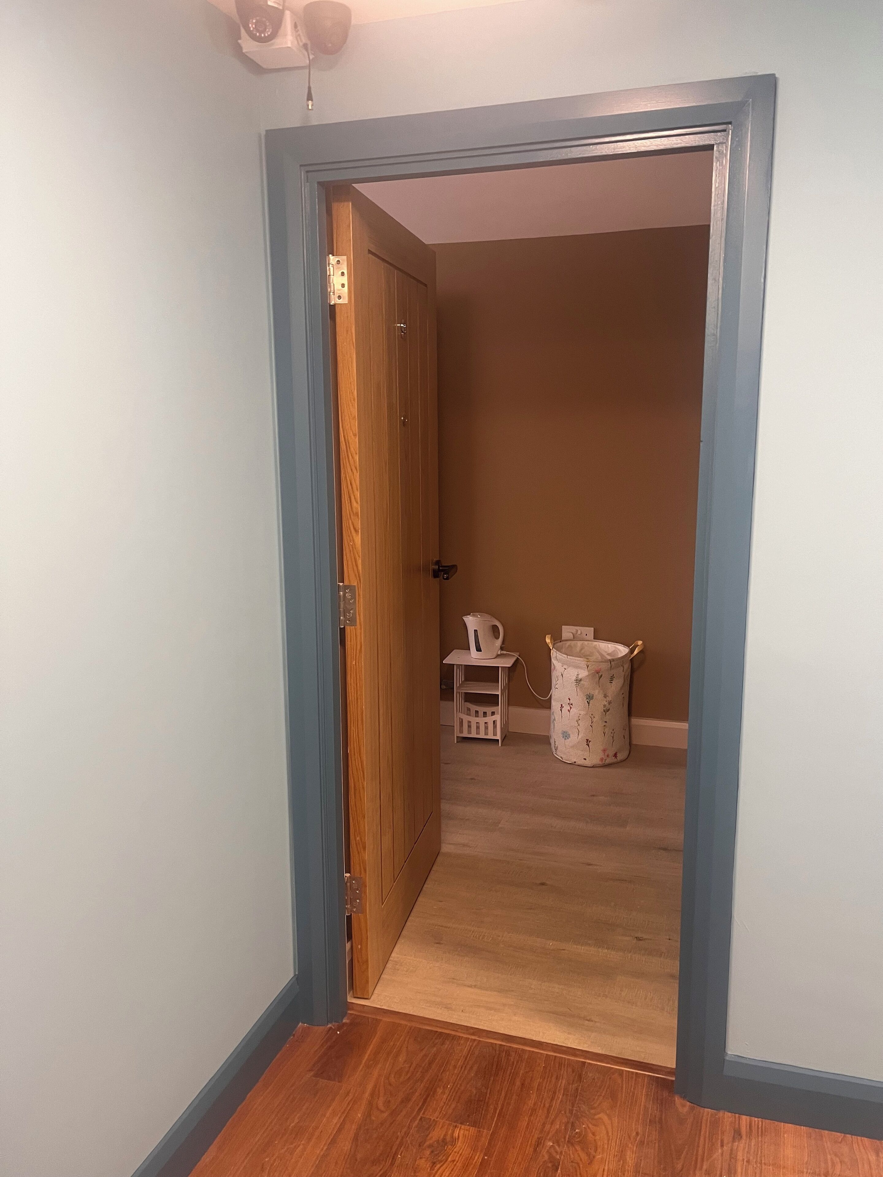 Un pasillo con una puerta de madera, una mesita con una tetera y un cubo de basura.