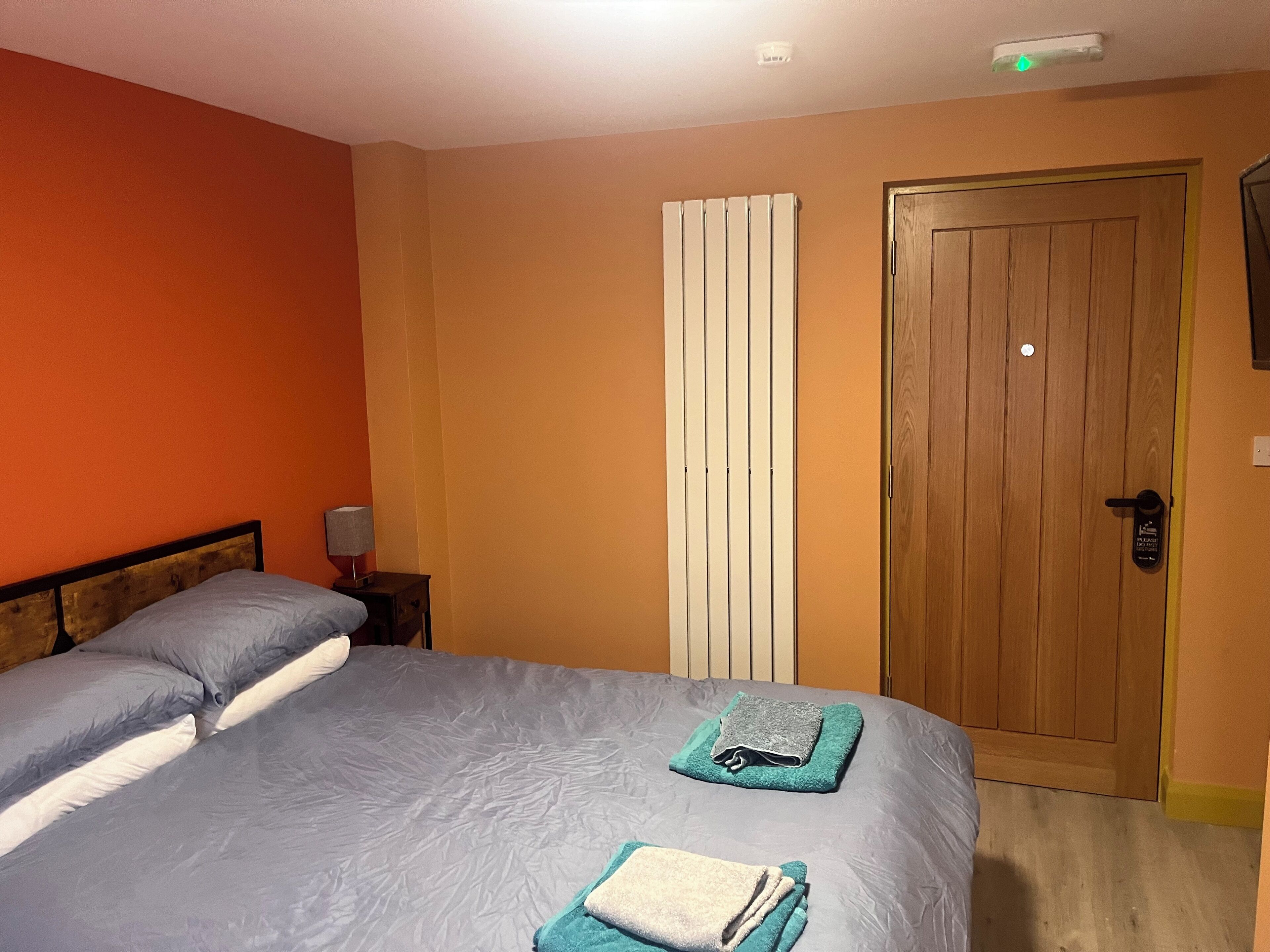 A bedroom with a bed, a nightstand, a lamp, a radiator, and a wooden door.