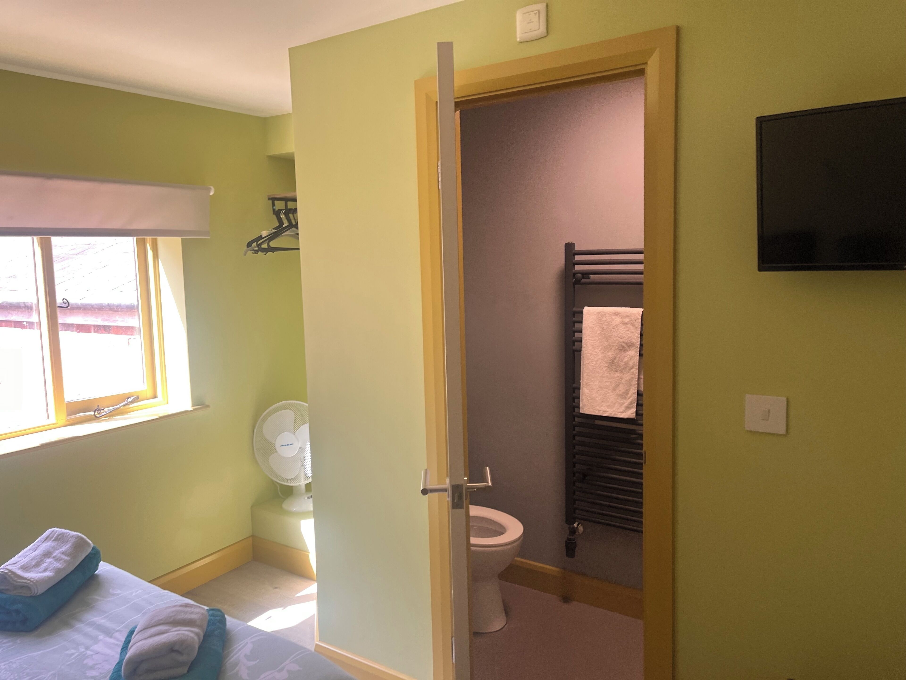 Una habitación con cama, ventana, ventilador, inodoro y perchero para toallas.