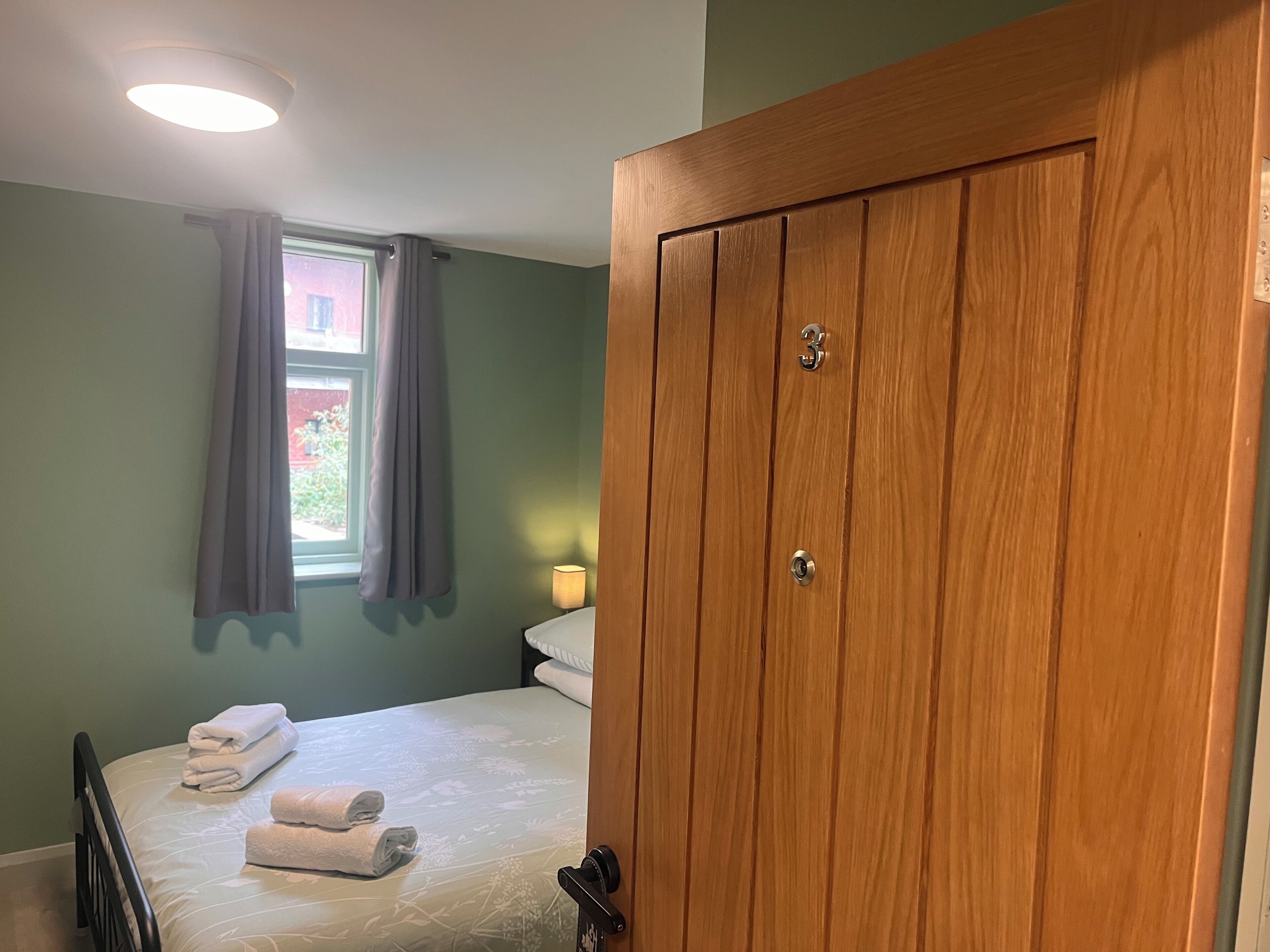 A bedroom with a wooden wardrobe, a bed with white linens, a window with curtains, and a bedside lamp.