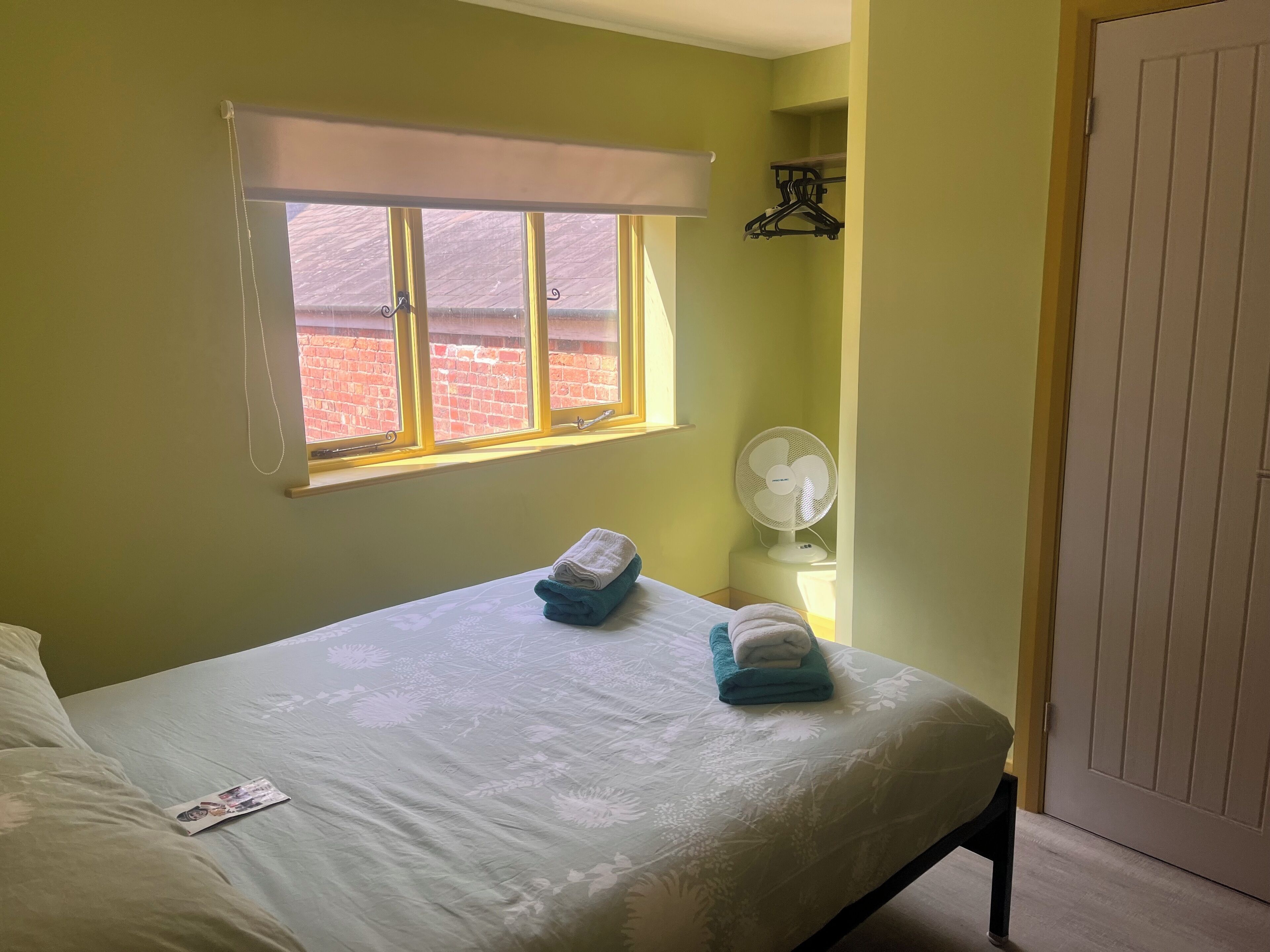 A room with a bed, a window with a view of a brick wall, a white fan, and a door.