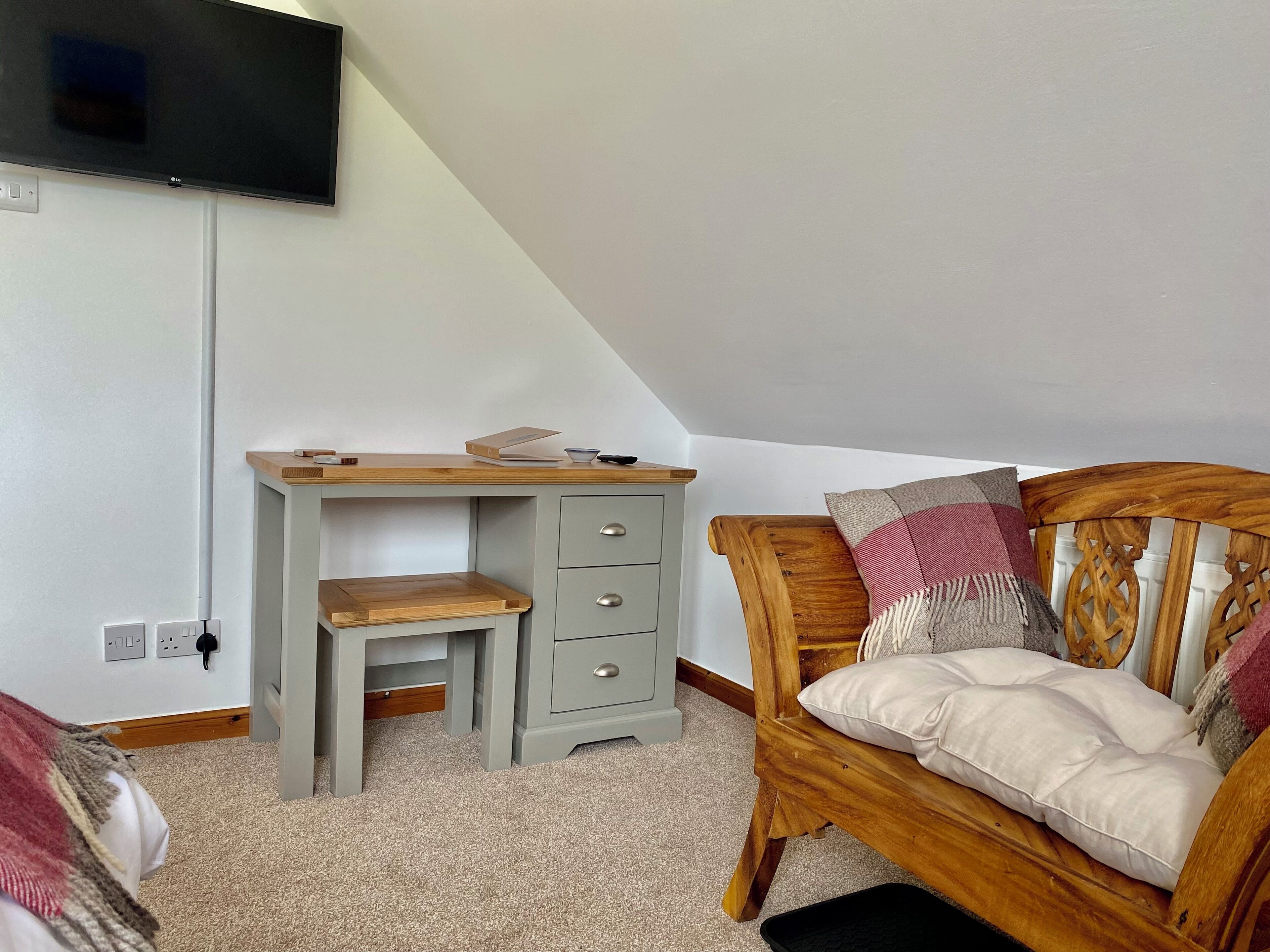 A bedroom with a wooden bed, a grey desk with drawers, a television mounted on the wall, and a carpeted floor.