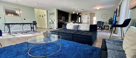 A modern living room with a blue shag rug, a glass coffee table, a black sectional sofa, and a dining area with a kitchen in the background.