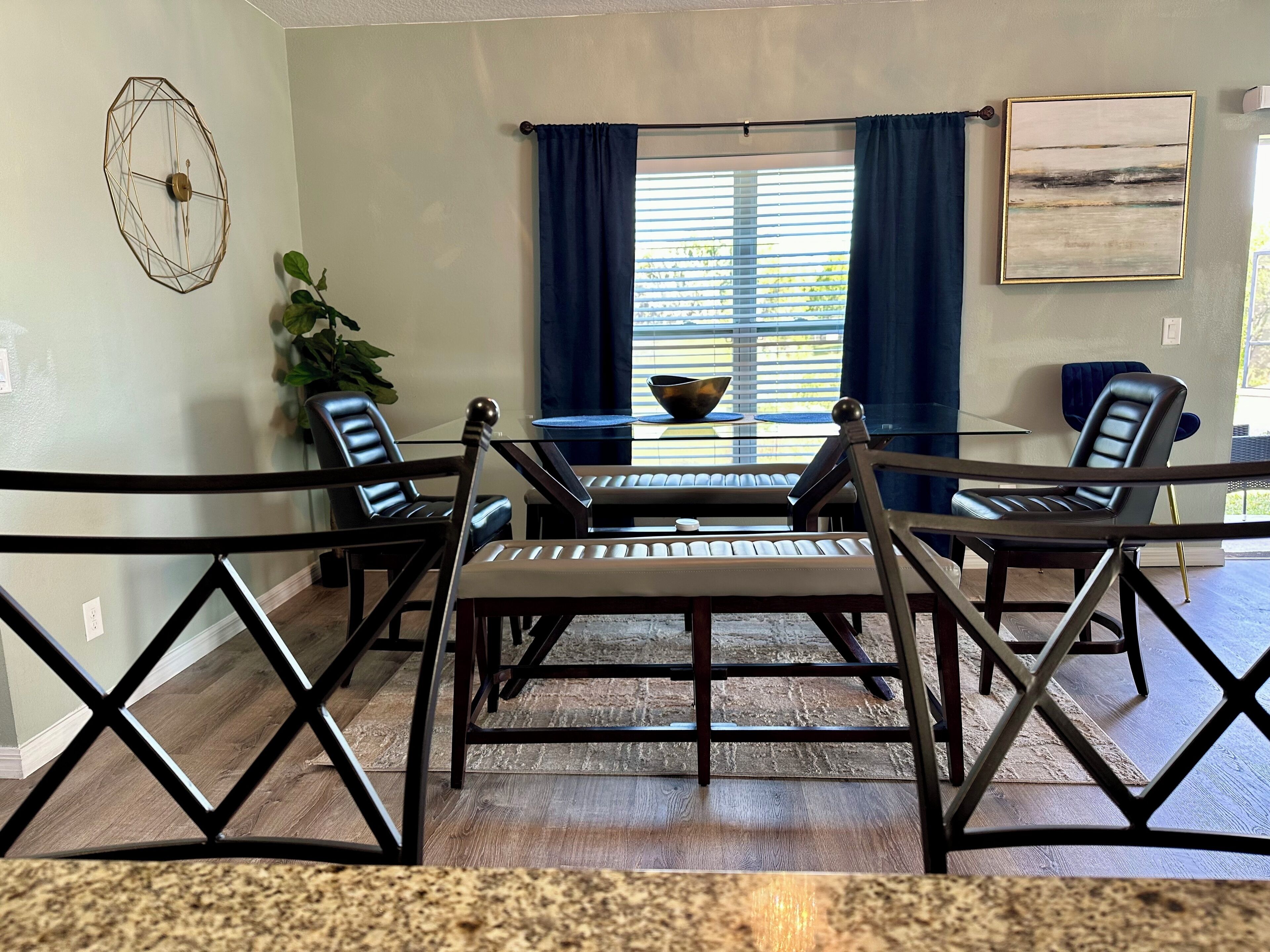 A dining area with a glass table, four chairs, a decorative wall clock, and a window with curtains.
