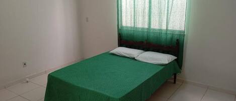 A single bed with a green bedspread and white pillows in a room with white walls and a window with green curtains.