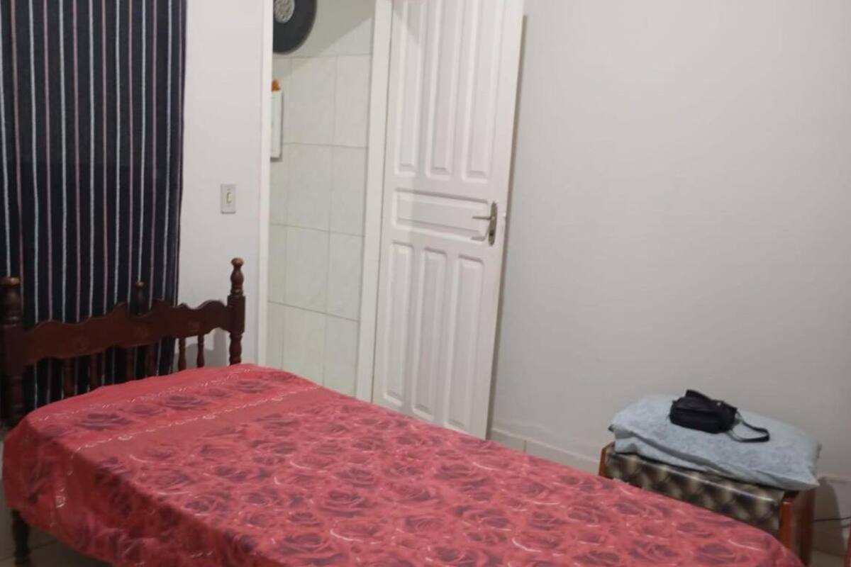 A small, neatly arranged bedroom with a single bed, a wooden chair, a small table with a bag, and a closed door.
