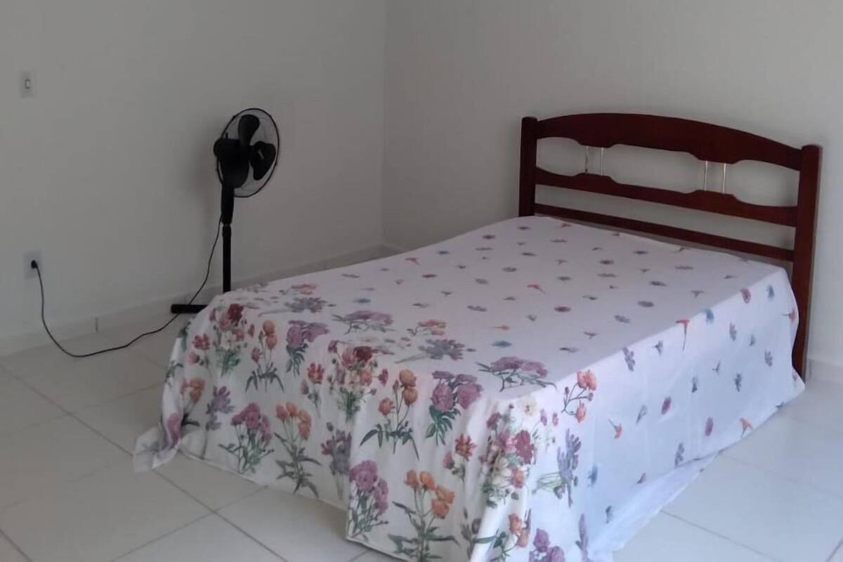A small, simple room with a single bed, a wall-mounted fan, and a plain white wall.