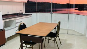 Een eetgedeelte met een tafel en stoelen, een keuken met witte kastjes, en uitzicht op een zonsondergang boven het water door een glazen wand.