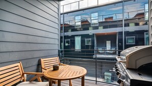 Een balkon met een houten tafel en stoelen, een barbecue en uitzicht op een modern gebouw met grote ramen.