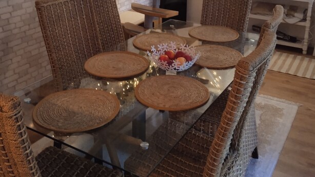 A dining area with a glass table surrounded by wicker chairs, a woven cat tree, and a decorative wall with ivy and string lights.