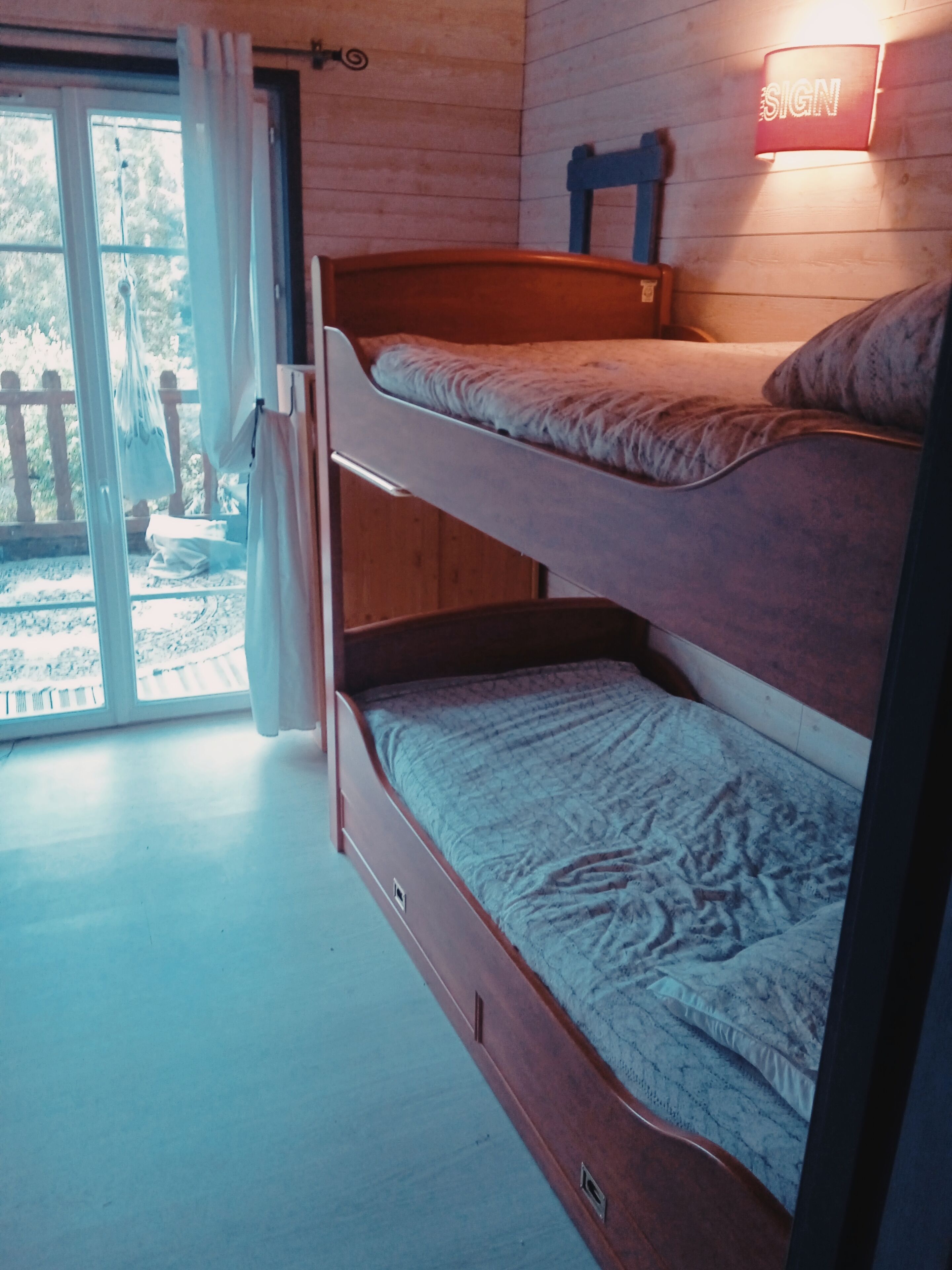 A bunk bed with a wooden frame and a lower bed with a patterned cover. There is a sliding glass door leading to an outdoor area with a wooden deck and a view of trees.