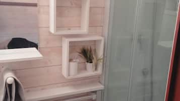 Une salle de bain avec un mur en bois, une cabine de douche en verre et des étagères blanches ornées d’une plante en pot.