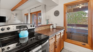 A kitchen with a black stove, a blue kettle, wooden cabinets, a clock, and a window with a view of trees. - Vista Serenity: Cozy Kiva, mountain views, 3 minutes from downtown (Santa Fe)