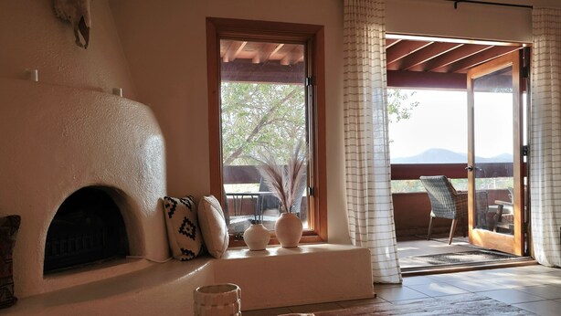 A cozy living room with a fireplace, a window with a view of the outdoors, and a wooden floor. - Vista Serenity: Cozy Kiva, mountain views, 3 minutes from downtown (Santa Fe)
