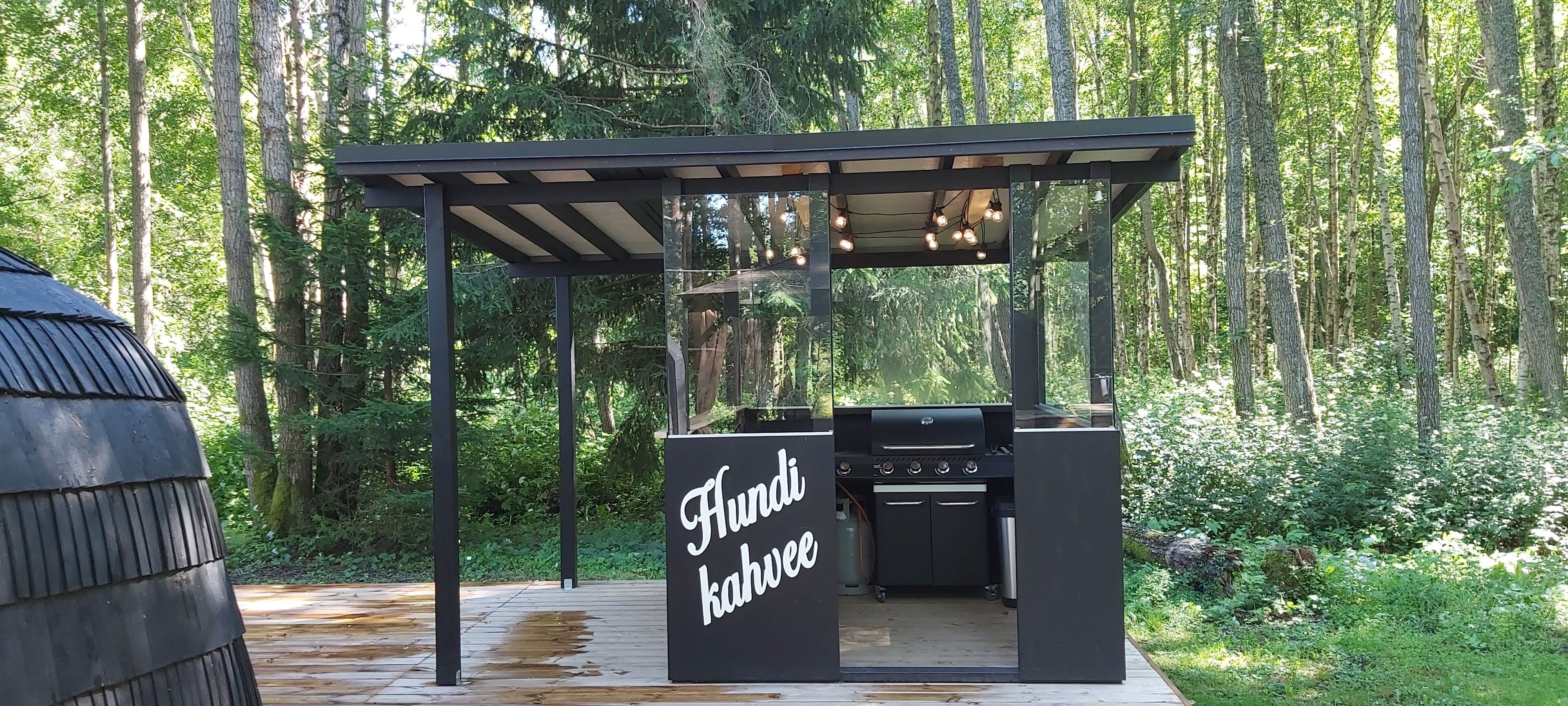 A modern outdoor barbecue area with a pergola and string lights, surrounded by a forest.