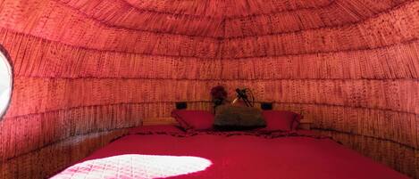 A cozy, circular room with a red bedspread and pillows, a small table with a vase, and a single window.