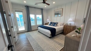 A modern bedroom with a bed, bedside tables, a ceiling fan, and a view of the outside. - Brand New Luxury Beach House - Hot Tub (Galveston)